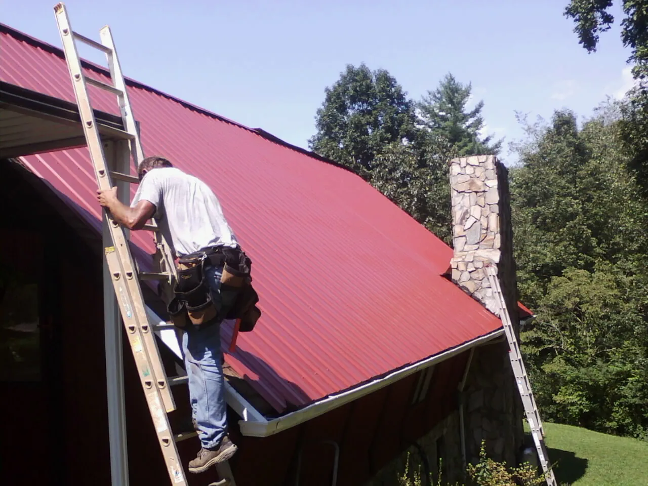 Roofer on a ladder beside a red metal roof and stone chimney — Soffit & Fascia Repair by Roofing Brothers in Stover